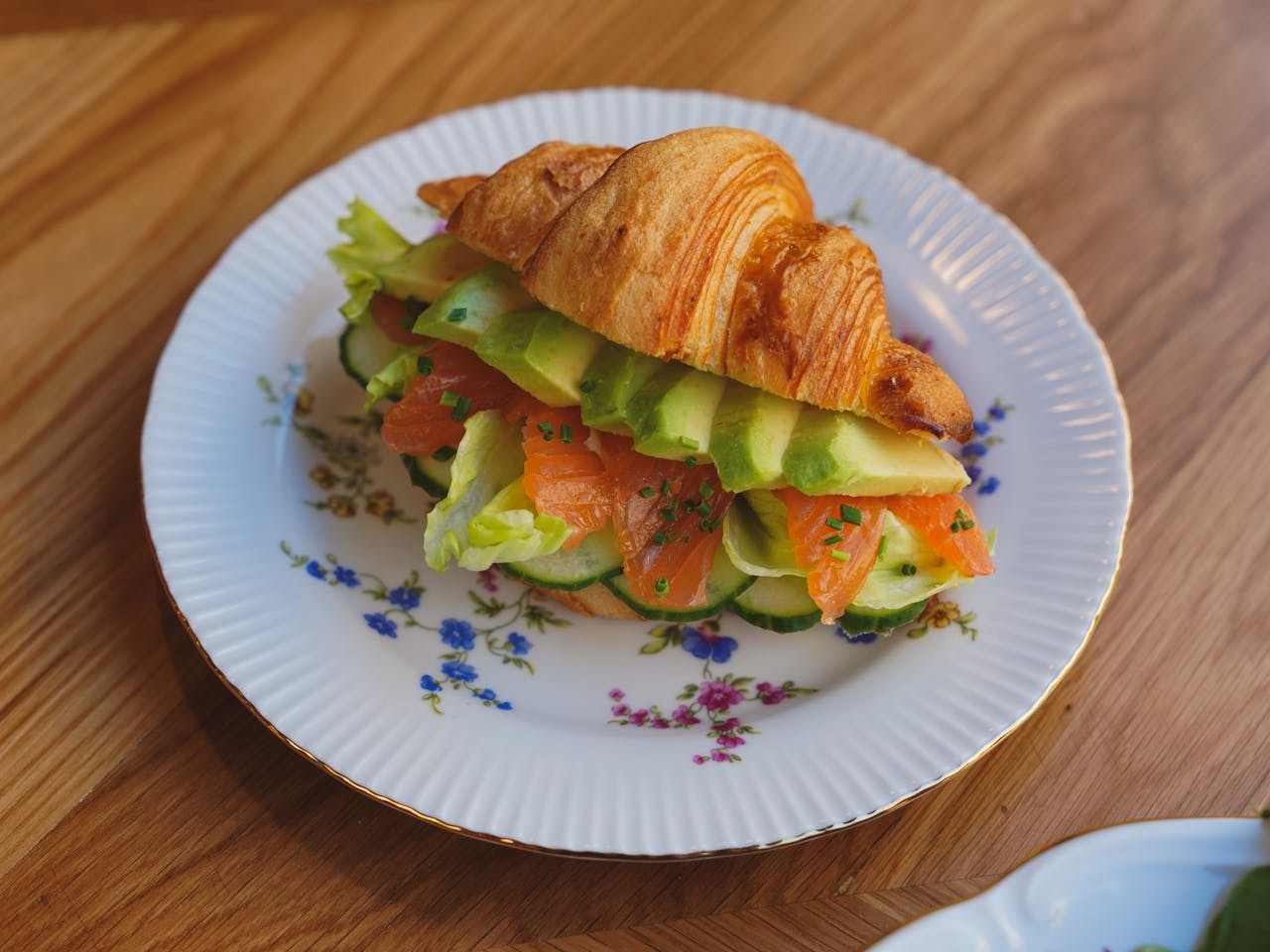 Tasty croissant sandwich filled with salmon, avocado, cucumber, and lettuce on a decorative plate.